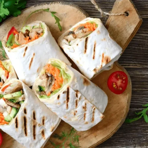 A wooden serving board featuring delicious grilled chicken wraps sliced in halves, showcasing a colorful filling of grilled chicken, fresh lettuce, shredded carrots, cucumbers, and a creamy sauce. Garnished with cherry tomatoes, lemon slices, and fresh herbs, this dish highlights the perfect grilled chicken wrap recipe for a healthy and flavorful meal.