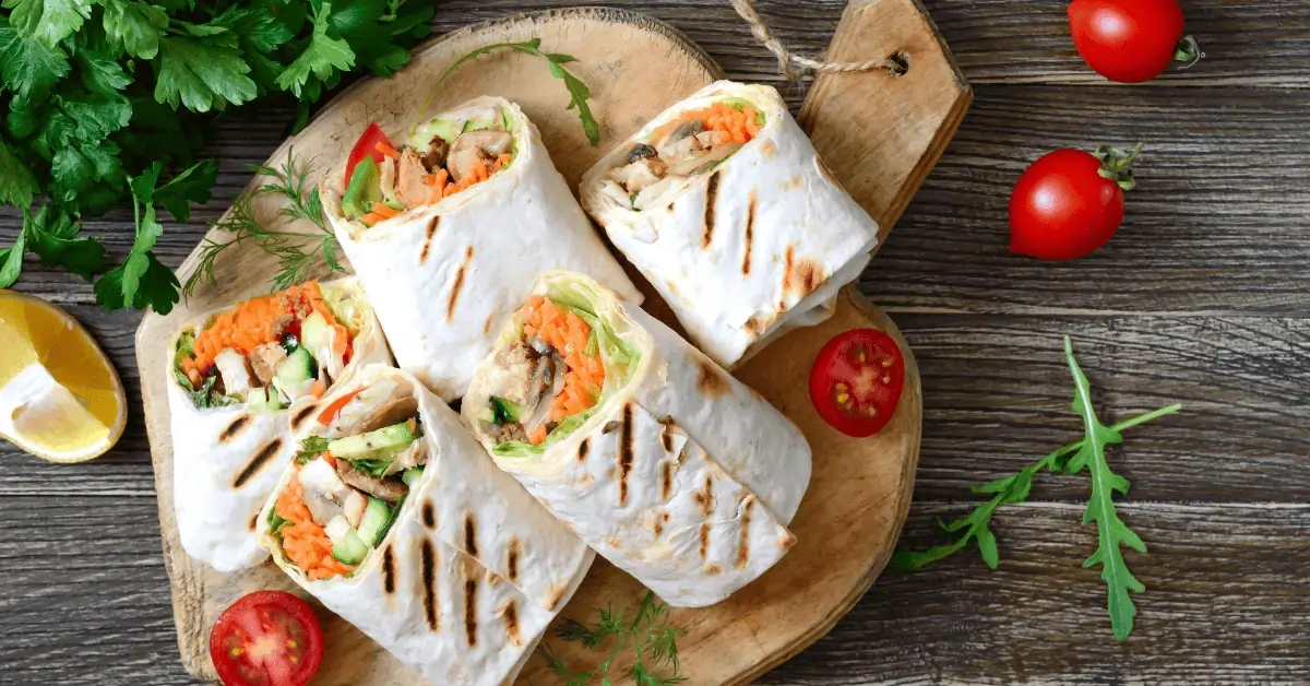 A wooden serving board featuring delicious grilled chicken wraps sliced in halves, showcasing a colorful filling of grilled chicken, fresh lettuce, shredded carrots, cucumbers, and a creamy sauce. Garnished with cherry tomatoes, lemon slices, and fresh herbs, this dish highlights the perfect grilled chicken wrap recipe for a healthy and flavorful meal.