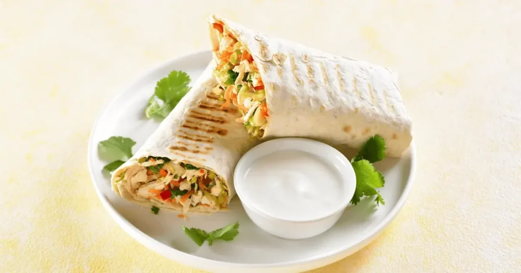 A perfectly grilled chicken wrap sliced open to reveal fresh vegetables and tender chicken filling, served with a creamy dipping sauce and garnished with cilantro on a white plate.