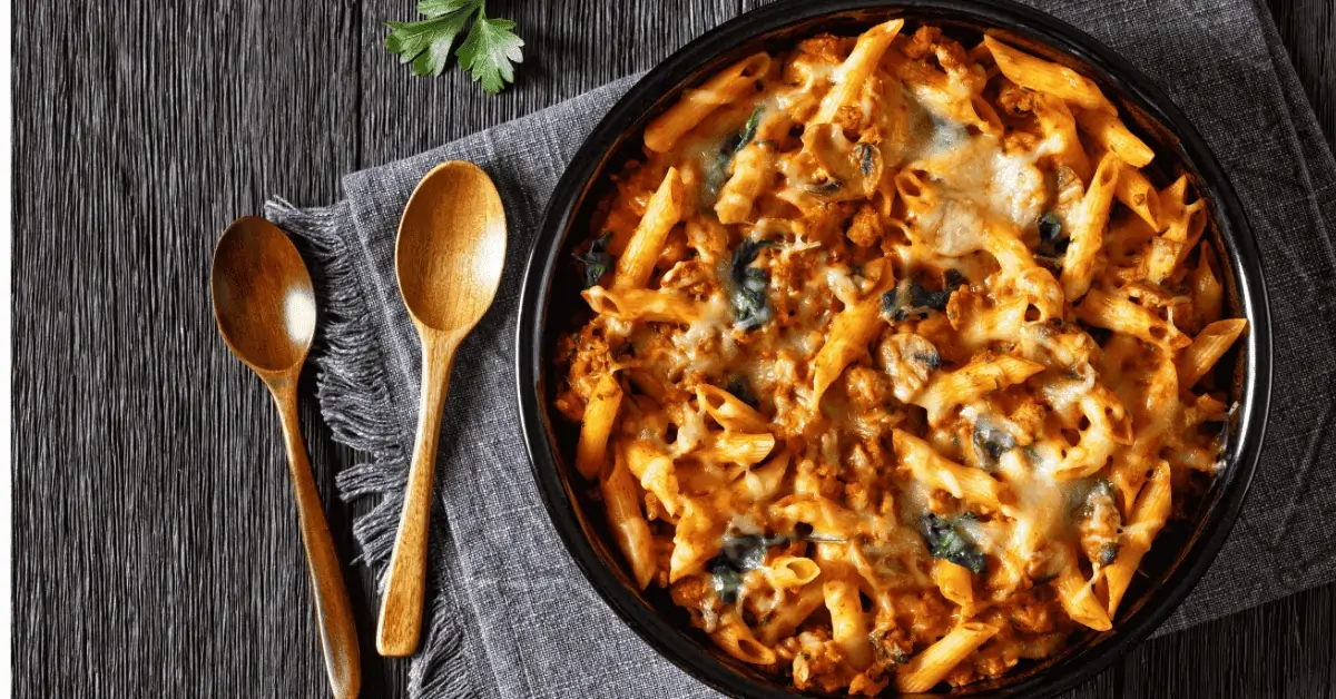 A freshly baked ground chicken pasta dish with tender penne, melted cheese, and spinach, served in a black baking dish with wooden spoons and garnished with fresh herbs.