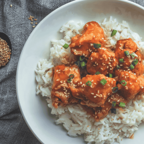A beautifully plated rice recipe with Korean chicken, garnished with sesame seeds and green onions, served on a white plate with sesame seeds sprinkled nearby.
