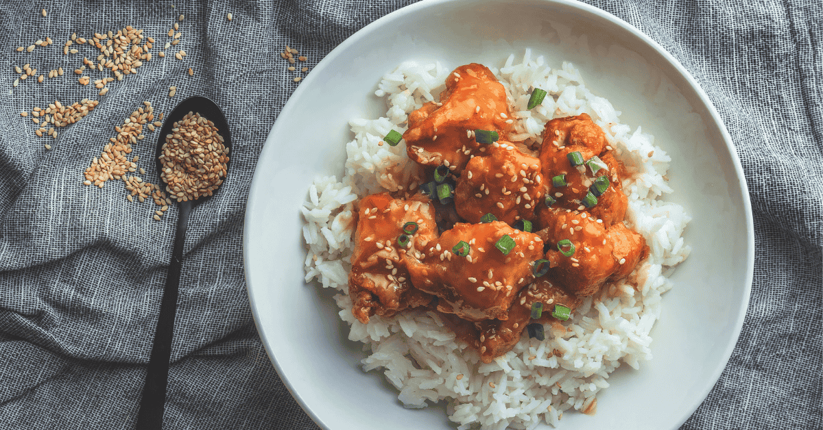 A beautifully plated rice recipe with Korean chicken, garnished with sesame seeds and green onions, served on a white plate with sesame seeds sprinkled nearby.