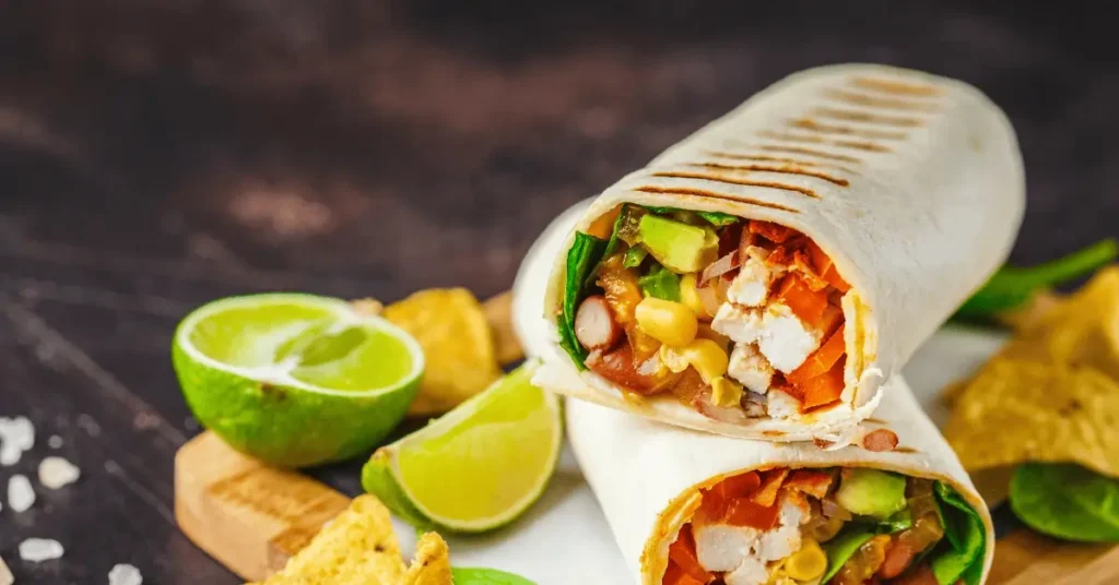 A chicken salad wrap recipe featuring a tortilla filled with grilled chicken, avocado, corn, and fresh vegetables, served with lime wedges and tortilla chips.